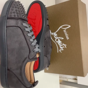 Christian Louboutin grå mocka sneakers - Snygga grå sneakers från Christian Louboutin i mocka med ikoniska röda sulor och coola nitar på tån. Skorna har snörning och en platt sula. Perfekta för dig som vill sticka ut med exklusiv design och edgy detaljer.  KVITTO FINNS