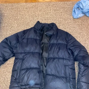 Svart pufferjacka från Everest - Svart pufferjacka från Everest med hög krage och dragkedja framtill. Jackan har en klassisk quiltad look och är perfekt för kalla dagar. Materialet är syntet och jackan har långa ärmar samt två fickor med dragkedja på sidorna. Storlek: S