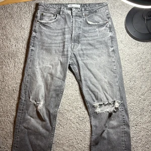 Grå slitna jeans från Zara - Grå jeans från Zara med snyggt slitna detaljer och hål på ena knät. Klassisk femficksmodell med raka ben och normal passform. Jeansen är tillverkade i bomull och har en cool, tvättad look som passar perfekt till streetwear. Dem är typ lite baggy så är inte min stil riktigt. Men dem har varit andvändna kanske 3 gånger sedan köp