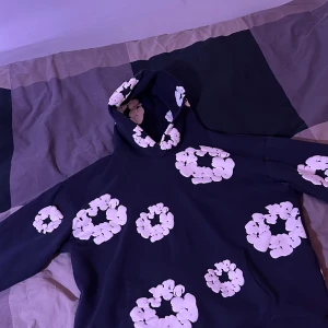 Svart hoodie med blommor från Denim Tears - Svart hoodie från Denim Tears med stora vita blommiga tryck över hela tröjan och huva. Tröjan har en klassisk känguruficka framtill och ribbade muddar vid ärmslut och nederkant. Perfekt för dig som vill sticka ut med en unik och trendig stil. Använd 2-3 gånger!!!!! Fortfarande i nyskick. Mörkblå!