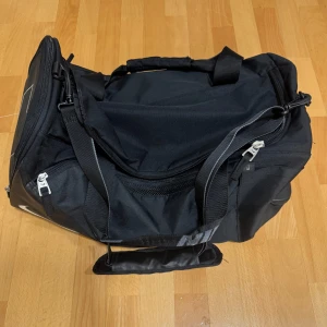 Svart sportbag från Nike - Stor svart sportbag från Nike med flera fack och dragkedjor. Väskan har rejäla handtag med vaddering och en justerbar axelrem. Snyggt Nike-tryck på sidan och detaljer i silver. Perfekt för gymmet eller träningen.