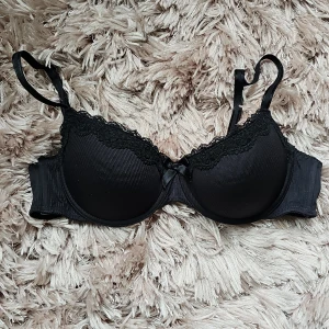 Svart spets-bh från Hunkemöller 80B - Snygg svart bh från Hunkemöller i storlek 80B. Bh:n har vadderade kupor med diskret ribbad struktur, spetsdetaljer längs kanten och en liten rosett framtill. Justerbara axelband och klassisk knäppning bak. Perfekt för dig som vill ha en stilren och feminin look.