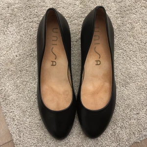 Svarta pumps från Unisa i skinn - Pris går att diskutera Stilrena svarta pumps från Unisa med rund tå och blockklack. Skorna är tillverkade i slätt skinn och har en klassisk, tidlös design som passar till många olika outfits. Perfekta för dig som gillar enkel och elegant stil.