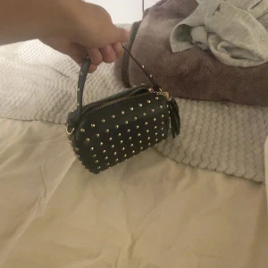 Svart handväska med nitar och tofs - Cool svart handväska i skinnimitation med guldiga nitar över hela väskan och en dekorativ tofs vid dragkedjan. Väskan har ett kort handtag med nitar och guldig dragkedja. Perfekt för dig som vill sticka ut med en edgy accessoar.