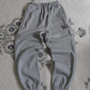 Gråa Stussy x Nike mjukisbyxor - Snygga grå mjukisbyxor från Stussy och Nike med broderad logga på vänster ben. Byxorna har elastisk midja med snörning, dragkedjeförsedda fickor och muddar vid bensluten. Perfekta för chill eller streetwear. Tillverkade i mjuk bomullsmix för maximal komfort.