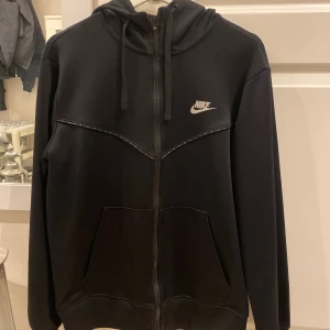 Svart Nike hoodie med dragkedja - Svart hoodie från Nike med hel dragkedja, huva med snörning och klassisk logga på bröstet. Storlek M. Använd mycket men fortfarande bra skick! Köparen står för frakt!