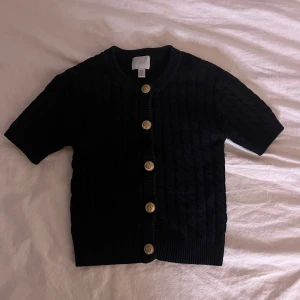 Svart kofta med guldknappar H&M - Kortärmad svart kofta från H&M med snygga guldknappar framtill. Tröjan är stickad med ett diskret kabelmönster och har ribbade kanter vid hals, ärmslut och nederkant. Perfekt för lager-på-lager och en clean look.