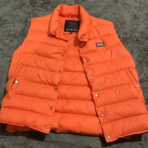 Orange dunväst från Cedrico - Säljer en riktigt snygg orange dunväst från Cedrico. Västen har quiltad design, hög krage och tryckknappar framtill. Perfekt för lager-på-lager och ger en cool pop av färg till din outfit. Innerficka med dragkedja och diskret logga på bröstet.