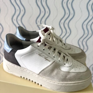 Axel Arigato Orbit Sneaker vit/blå/röd - Snygga Axel Arigato Orbit sneakers i vitt skinn och mocka med blå och röda detaljer. Klassisk siluett med rund tå, snörning och platt sula. Märkeslogga i guld på sidan och coolt mönstrad yttersula. Perfekta för dig som gillar stilrena och trendiga sneakers.