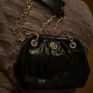 Svart axelväska från Tory Burch i skinn - Snygg svart axelväska från Tory Burch i glansigt skinn med guldfärgade kedjor och detaljer. Väskan har dragkedja upptill, innerfack med dragkedja och ikonisk logga framtill. Perfekt storlek för det viktigaste och ger en lyxig känsla.