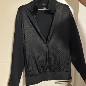 Svart bomberjacka från BOSS äkta (priset är inte hugget i sten )  - Svart bomberjacka från BOSS med diskret mönster på framsidan och hel dragkedja. Jackan har ribbade muddar och krage, samt långärmad design. Materialet är merceriserad bomull som ger en lyxig känsla och snygg finish. Perfekt för dig som gillar stilrena detaljer.