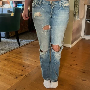 Ljusblå slitna jeans - Säljer ett par ljusblå jeans med utsvängda ben och flera slitningar och hål på knäna,låg midja och är tillverkade i bomullsdenim. Perfekta för en avslappnad och trendig look.