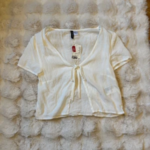 Vit croppad blus med knyt från H&M - Superfin vit croppad blus från H&M med korta ärmar och v-ringning. Toppen har en snygg knytning framtill och är gjord i ett lätt, luftigt bomullstyg. Perfekt för varma dagar och enkel att styla med jeans eller kjol.