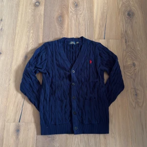 Mörkblå kofta från Polo Ralph Lauren - Snygg mörkblå kofta från Polo Ralph Lauren med kabelstickat mönster och röd broderad logga på bröstet. Koftan har v-ringning, knappar framtill och två fickor. OBS: storlek L men sitter som S. 