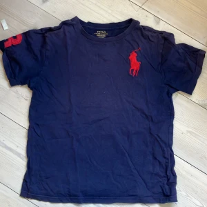 Marinblå t-shirt från Polo Ralph Lauren, strl 10-12 - Snygg marinblå t-shirt från Polo Ralph Lauren med stor röd logga broderad på bröstet och siffra på ärmen. Klassisk rund hals och korta ärmar. Tillverkad i mjuk bomull för skön känsla hela dagen.