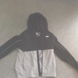 Svart och grå vindjacka från McKenzie - Snygg tvåfärgad vindjacka från McKenzie med svart överdel och grå nederdel. Jackan har huva med snörning, dragkedja framtill och praktiska fickor. Perfekt för blåsiga dagar och enkel att matcha med streetwear.