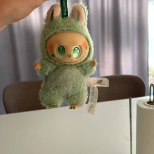 The Monsters Plush Doll - Söt Pop Mart The Monsters plush-docka i grönt lurvigt tyg med stora gröna ögon och charmigt leende. Dockan är i gott skick men har lite slitage och färgskiftningar baktill. Perfekt för samlare eller som present till Pop Mart-fans.