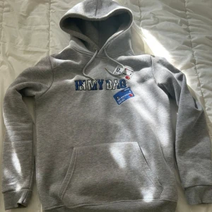 Grå hoodie IN MY BAG med brodyr - Grå hoodie från IN MY BAG med broderad text och coola kreditkortsdetaljer på bröstet. Tröjan har huva med snörning, magficka och ribbade muddar. Materialet är mjuk bomullsblandning och passformen är normal. Perfekt för chill dagar eller streetwear-stil.