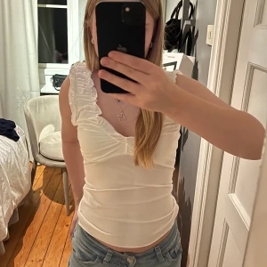 Vit volangtopp med v-ringning - Supersöt vit topp med volangdetaljer längs axelbanden och djup v-ringning. Figurnära passform och ärmlös design. Prislappen sitter kvar och har aldrig använts, banden är väldigt långa dock. 