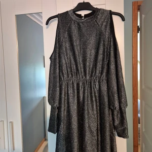 Glittrig svart kortklänning med cut out - Supersnygg svart glittrig kortklänning med markerad midja och långa ärmar med trendiga cut out-detaljer på axlarna. Klänningen har rund halsringning och ett lätt glansigt material som verkligen sticker ut. Perfekt för dig som vill ha en cool och festlig look.