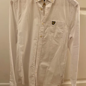 Vit skjorta från Lyle & Scott - Klassisk vit skjorta från Lyle & Scott med lång ärm, bröstficka och den ikoniska gula örnloggan. Skjortan har knappar hela vägen och är tillverkad i mjuk bomull. Perfekt för en clean och stilren look.