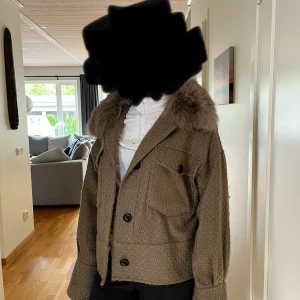 Beige jacka med päls från Meotine - Säljer en beige jacka från Meotine med snyggt diskret mönster och stor fluffig päls runt kragen. Jackan har stora knappar framtill, två bröstfickor och breda ärmslut med knappdetalj. Boxig och lite croppad passform, perfekt för höst och vinter.