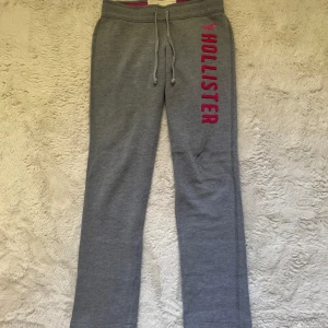 Grå mjukisbyxor från Hollister - Grå mjukisbyxor från Hollister med rosa logga längs benet. Byxorna har dragsko i midjan och är fodrade med mjukt tyg på insidan. Perfekta för chill dagar och har en avslappnad passform.