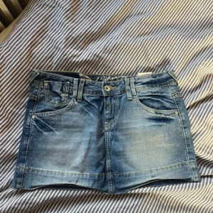 Blå jeanskjol från Cron-X, storlek 28 - Snygg blå jeanskjol från Cron-X i klassisk femficksmodell med bälteshällor och knappstängning. Kjolen är tillverkad i 100% bomull och har slitningar för en cool look. Passar perfekt till sneakers eller boots och har en normal passform.