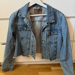 Croppad jeansjacka med nitar från Amisu - Snygg croppad jeansjacka från Amisu i ljusblå denim. Jackan har silverfärgade nitar på krage och bröstfickor och vanliga fickor, rå kant nertill och klassiska knappar framtill. Perfekt för dig som vill ha en edgy och trendig look.😊