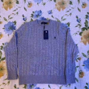 Ljusgrå kabelstickad tröja Polo Ralph Lauren - Kabelstickad ljusgrå tröja från Polo Ralph Lauren med klassisk rund halsringning och broderad logga på bröstet. Tröjan har ribbade muddar och är tillverkad i mjuk ullblandning. Perfekt för lager-på-lager och snygg till jeans.