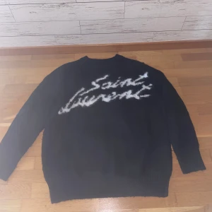 Svart stickad Saint Laurent-tröja - Svart stickad tröja från Saint Laurent med vit logotyp i graffiti-stil över bröstet. Tröjan har rund halsringning, långa ärmar och ribbade muddar. Perfekt för dig som vill ha en lyxig och cool statement-piece i garderoben.