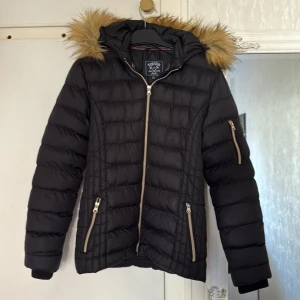 Svart pufferjacka från Vinson Polo Club - Svart pufferjacka från Vinson Polo Club med snygg fuskpäls på huvan och guldiga dragkedjor. Jackan har quiltad design, tre fickor med dragkedja och badge på ärmen. Perfekt för kalla dagar och riktigt stilren look.