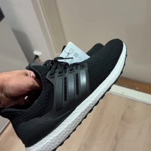 Adidas Ultraboost svarta sneakers - Svarta Adidas Ultraboost sneakers med stickad ovandel och klassiska tre ränder på sidan. Skorna har en vit, responsiv Boost-mellansula och svart yttersula med greppvänligt mönster. Perfekta för dig som gillar sportig stil och komfort. Snörning framtill och rund tå.