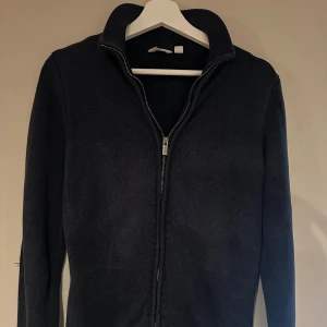 Mörkblå stickad kofta från Dobber - Mörkblå stickad kofta från Dobber med hel dragkedja framtill och ribbad krage. Klassisk och enkel design med långärmad passform, perfekt för lager-på-lager-stil under kyliga dagar.