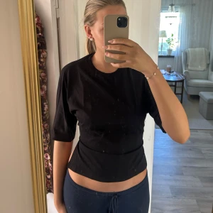 Svart Nelly topp med knyt i ryggen - Svart croppad topp i mjuk bomull med halvlånga ärmar och rund halsringning. Baksidan har en öppen rygg med bred knytning som ger en snygg detalj. Perfekt för dig som gillar trendiga och stilrena plagg.