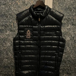 Exklusiv Morris dunväst (M) - Exklusiv dunväst från Morris, en ikonisk detalj i varje stilmedveten garderob. Den svarta färgen tillsammans med den eleganta brodyren på bröstet och den diskreta logotypen på ryggen ger västen en sofistikerad och tidlös look.  Tillverkad i högkvalitativa material med lätt fyllning som ger värme utan att kännas tung. Designad med hög krage, praktiska sidfickor med dragkedja och en bröstficka som förstärker den stilrena silhuetten. Perfekt att bära över en stickad tröja, skjorta eller hoodie.