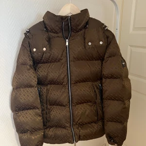 Brun pufferjacka från BOSS - Säljer en brun pufferjacka från BOSS med diskret rutmönster och hög krage. Jackan har dragkedja framtill, två fickor med dragkedja och snygga metalldetaljer vid axlarna. Märket syns tydligt på ärmen och insidan. Perfekt för kalla dagar.