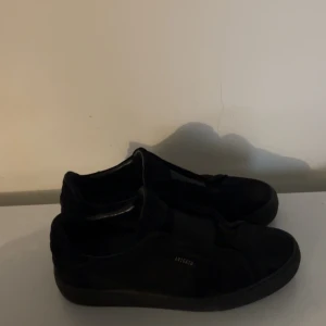 Svarta Arigato sneakers i mocka - Säljer ett par svarta sneakers från Axel Arigato med stilren design och låg profil. Skorna är tillverkade i mjuk mocka med platt sula och rund tå. Diskret Arigato-logga i guld på sidan. Perfekta för dig som gillar minimalistisk stil. I storlek 45, pris kan diskuteras så tveka inte på att höra av er.