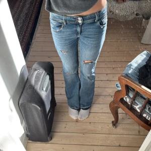 Blå bootcut jeans med slitningar - Säljer ett par blå jeans med bootcut passform och slitningar på båda benen. Jeansen har låg midja, klassiska fem fickor och dubbla knappar framtill. Materialet är denim och färgen är mellanblå med lätt tvättad look. Dem är i st 164 men passar mig som är 175 cm❤️