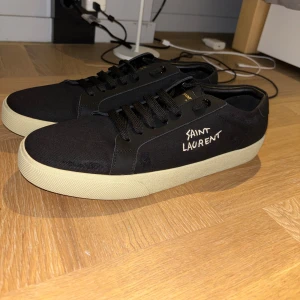 Saint Laurent skor - Svarta sneakers från Saint Laurent med vit sula och handskriven logga på sidan. Skorna har snörning, ovandel i textil och detaljer i skinn. Klassisk låg modell med rund tå och diskret branding på häl och sula. Perfekta för en clean och stilren look.