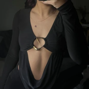 Långärmad topp med öppen front - En svart långärmad topp med djup v-ringning och öppen front. Toppen har en snygg metallring i guld mitt fram som håller ihop tyget och ger en unik look. 