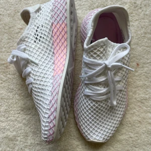 Adidas Deerupt Runner vita/rosa sneakers - Säljer ett par limiter edition Adidas Deerupt Runner sneakers i vitt med rosa detaljer. Skorna har ett unikt nätmönster över hela ovandelen och sula, samt klassisk snörning. Insidan är ljusrosa och materialet är lätt och andas, perfekt för en sportig och trendig look. Oanvända. 