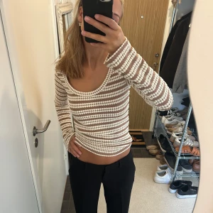 Randig croppad långärmad topp - Säljer en snygg croppad långärmad topp med vita och beige ränder. Toppen har en rundad urringning och är stickad i ett genombrutet mönster som ger en cool och trendig look. Perfekt för dig som vill sticka ut med en unik stil.