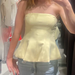 Gul korsett-topp med volangkant - Superfin ljusgul korsett-topp med axelbandslös design och utsvängd volangkant nedtill. Sitter tight upptill och ger en snygg siluett. Perfekt att styla med jeans eller kjol för en trendig look.