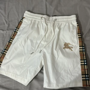 Vita Burberry shorts med rutiga sidor - Snygga vita shorts från Burberry med klassiska rutiga paneler längs sidorna och guldfärgat Burberry-ryttaretryck på ena benet. Elastisk midja med snörning för skön passform.  