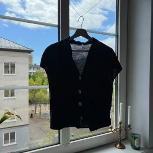 Svart linnetopp med knappar från MNG - Svart, luftig linnetopp från MNG med korta ärmar och v-ringning. Toppen har tre stora knappar framtill och är tillverkad i ett tunt, svalt linnematerial. Perfekt för dig som gillar minimalistisk och stilren look.