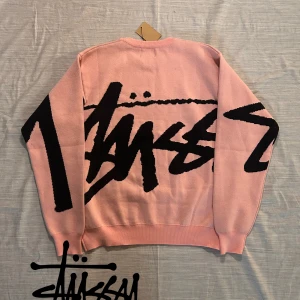 Rosa stickad tröja från Stüssy XL - Säljer en rosa stickad tröja från Stüssy i storlek XL. Tröjan har ett stort svart grafiskt tryck över hela framsidan och ärmarna. Rund halsringning och ribbade muddar. Perfekt för dig som gillar streetwear och vill sticka ut.