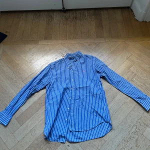 Blå randig skjorta från Ralph Lauren - Snygg blå skjorta från Ralph Lauren med vita vertikala ränder och klassisk krage. Skjortan har långa ärmar, knappar framtill och den ikoniska rosa loggan broderad på bröstet. Perfekt för en clean och stilren look.