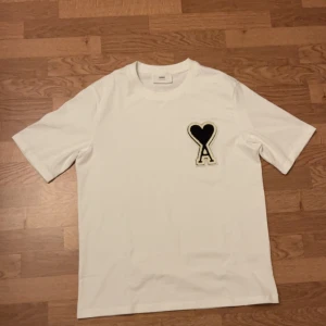 Vit t-shirt från Ami hearts med hjärtdetalj - Vit t-shirt från Ami hears med svart hjärta och bokstaven A broderat på bröstet. Klassisk passform, rund hals och korta ärmar. Tillverkad i mjuk bomull för en clean och stilren look. Storleken är S men passar M. PRIS KAN DISKUTERAS!