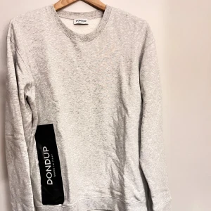 Grå sweatshirt från Dondup i bomull - Ljusgrå sweatshirt från Dondup med svart logga på sidan. Tröjan har rund halsringning och långa ärmar. Tillverkad i mjuk bomull, perfekt för en chill och stilren look.